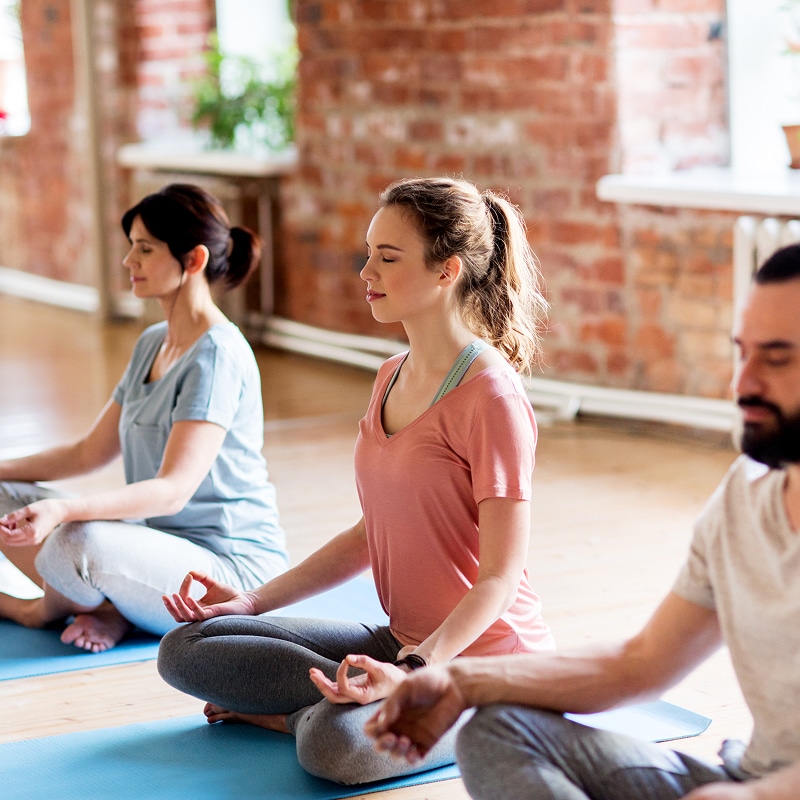 Trois personnes méditent calmement, assises en lotus sur des tapis de yoga bleus, les yeux fermés et mains en mudra, dans un studio chaleureux.