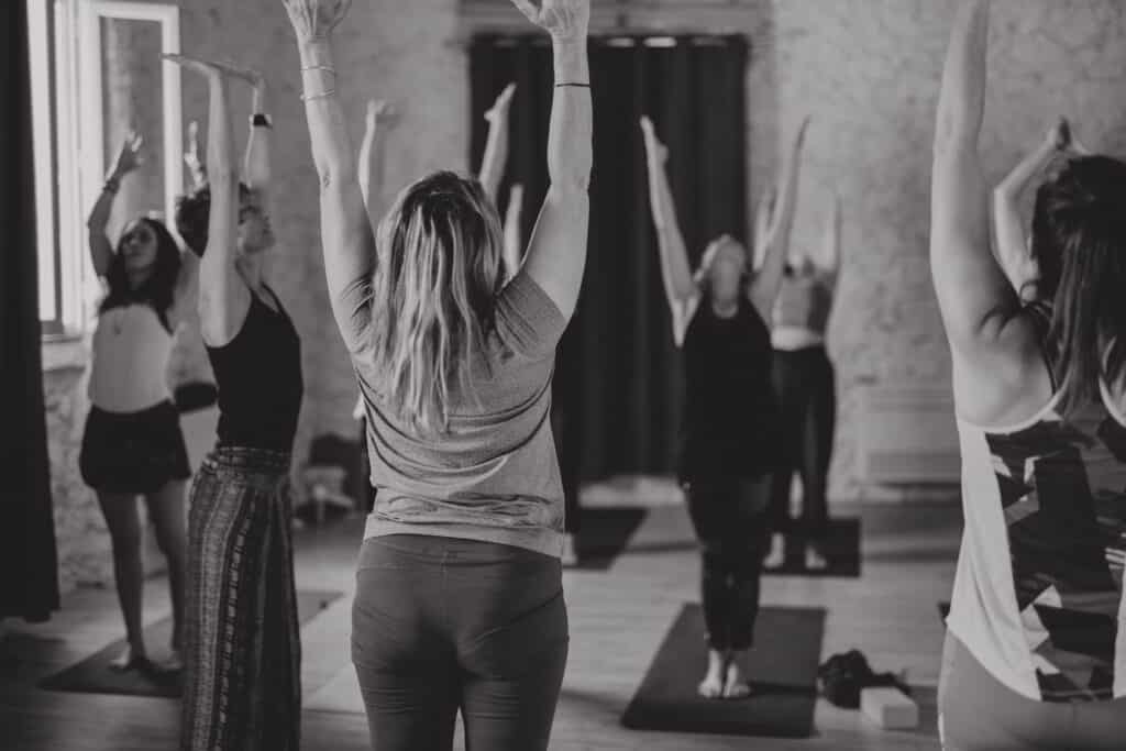 Groupe de femmes en cours de yoga, vue de dos, bras levés vers le haut. Scène de studio en noir et blanc.