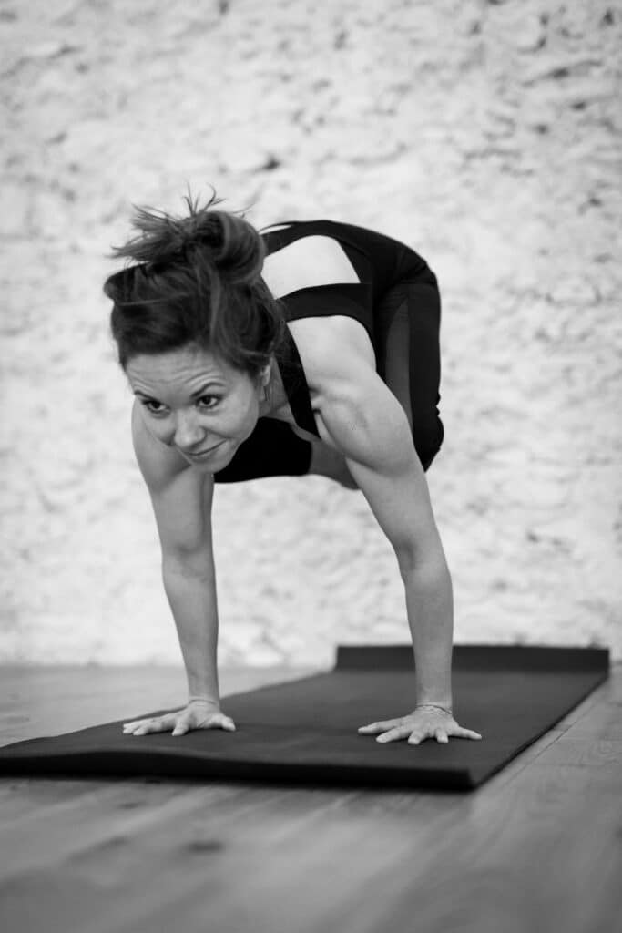 Femme en Bakasana (pose du corbeau) sur un tapis de yoga noir, en équilibre sur les mains, avec un léger sourire. Image noir et blanc.