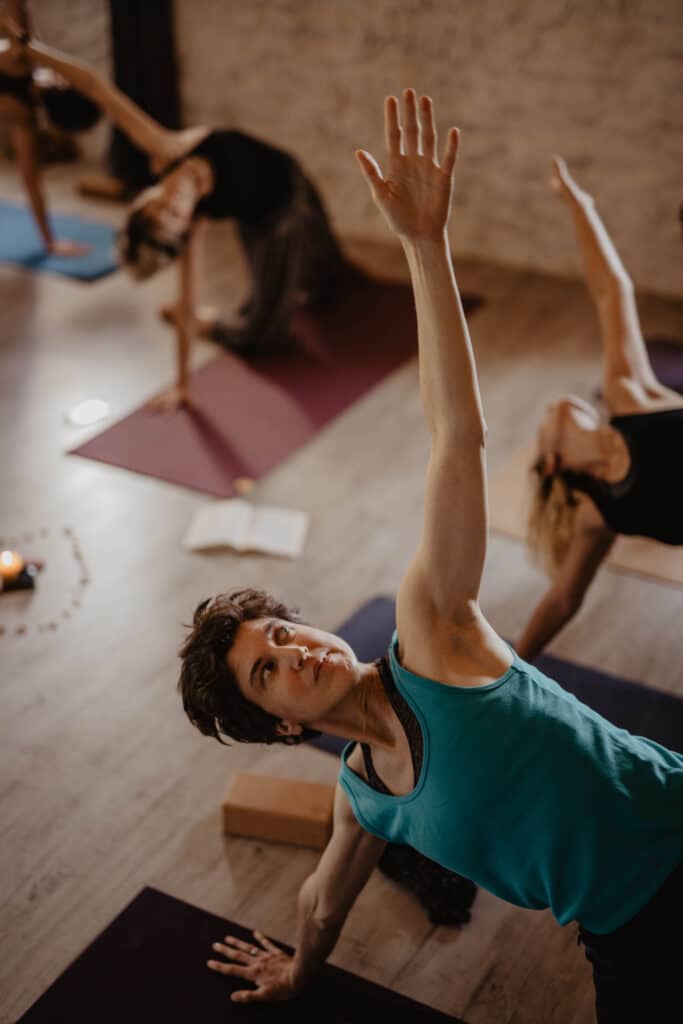 Femme en top turquoise pratiquant une pose de yoga latérale, bras levé, le regard concentré, dans un studio avec d'autres yogis.