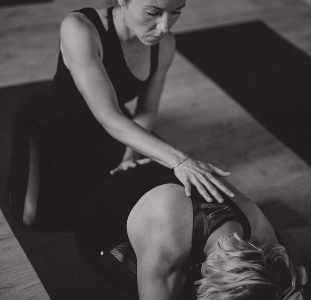 Professeure de yoga en noir guidant une élève en posture de relaxation, main sur le dos. Ambiance monochrome, calme et concentrée.