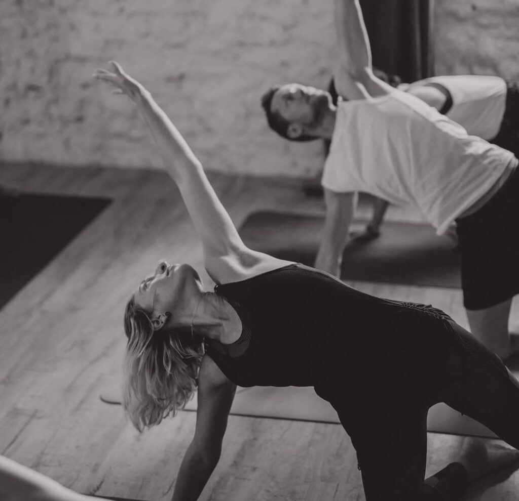 Femme et homme pratiquant le yoga en studio. La femme réalise une pose d'étirement du dos, bras levé. Un homme exécute une pose similaire.