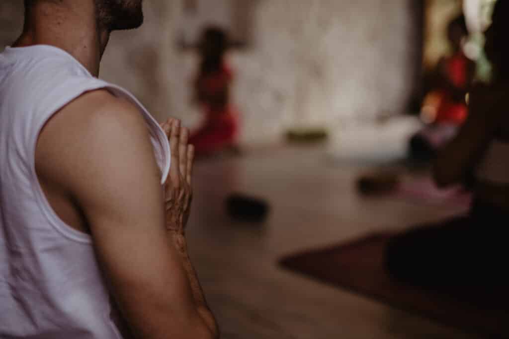 Homme en t-shirt blanc, mains en Namaste, méditant. Assis sur un tapis de yoga, avec d'autres participants flous en arrière-plan.