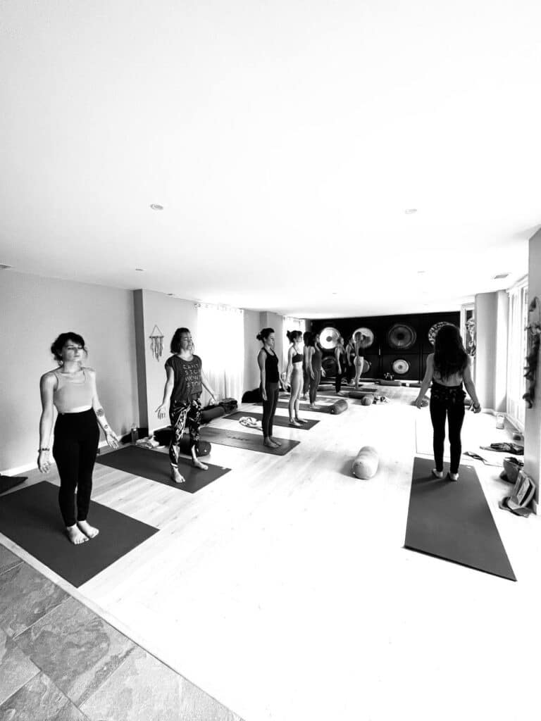 Sept femmes sur tapis de yoga dans un studio lumineux avec gongs muraux. Une femme porte un t-shirt "CAN'T STOP WON'T STOP".