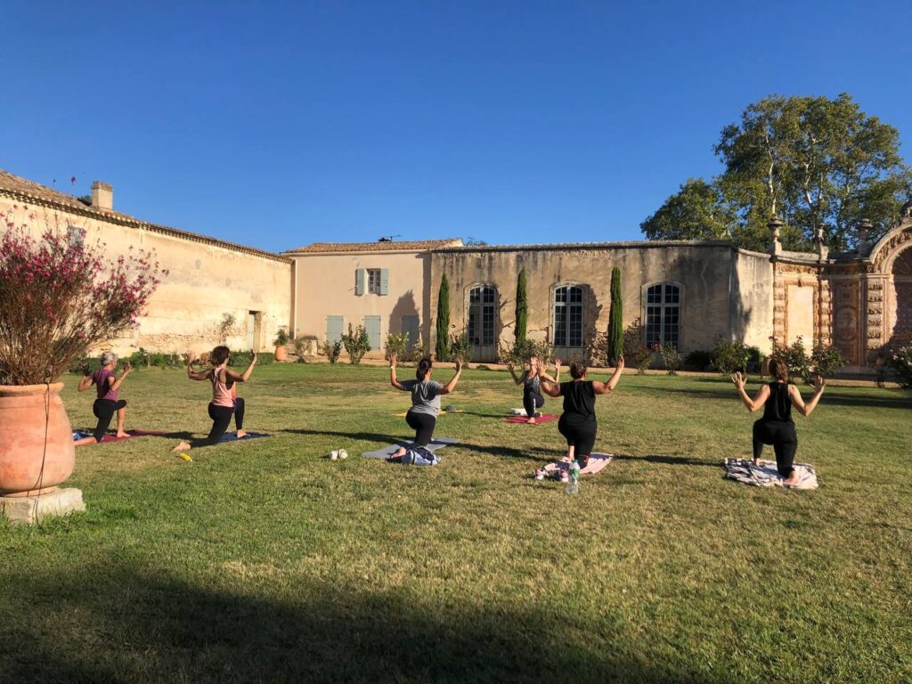 Femmes pratiquant le yoga en extérieur sur une pelouse verte. Elles sont en pose de fente, bras levés, devant un mas traditionnel sous ciel bleu.