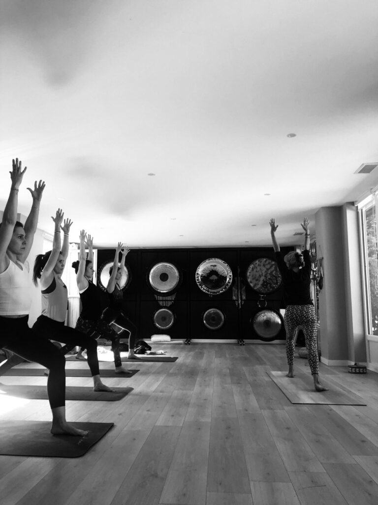 Groupe de personnes en pose de yoga bras levés dans un studio lumineux. Une instructrice mène la séance avec des gongs en arrière-plan.