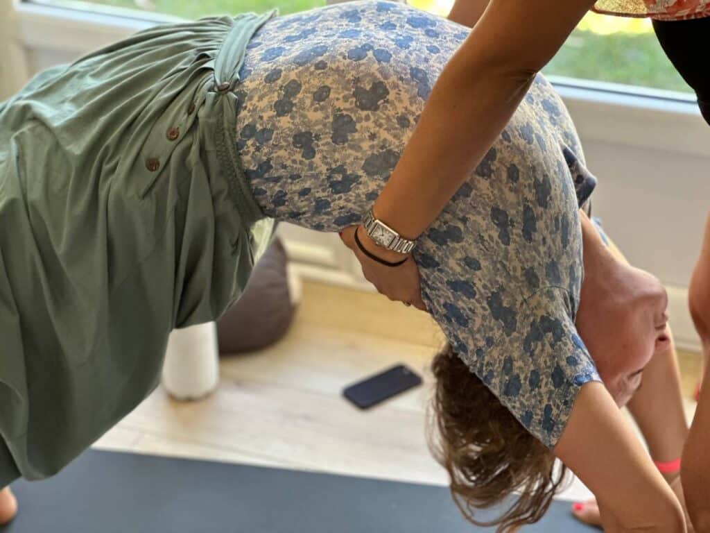 Personne en yoga effectuant une profonde flexion arrière (pont) assistée. Sur tapis noir, vêtue de fleurs bleues et vert kaki.